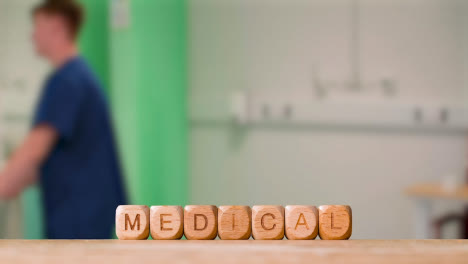 Concept-With-Wooden-Letter-Cubes-Or-Dice-Spelling-Medical-Against-Background-Of-Nurse-Wheeling-Patient-In-Hospital-Bed